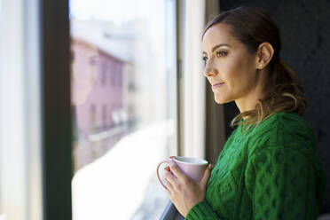 Frau hält Kaffeetasse, während sie zu Hause am Fenster steht - JSMF01748