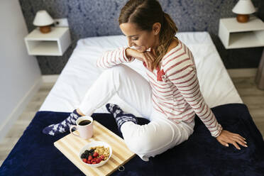 Smiling woman looking at food and coffee while sitting on bed at home - JSMF01750