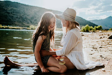 Mutter und Tochter lächelnd und kuschelnd am Seeufer an einem sonnigen Tag - CAVF89491