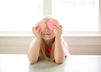 Cute blond girl holding pink frosted sugar cookies over eyes - CAVF89505