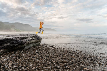 Junges Kind mit lockigem Haar springt von einem Felsen am Strand in Neuseeland - CAVF89520