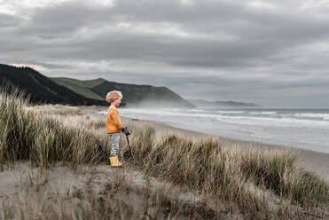 Blonder Junge mit lockigen Haaren beobachtet das Meer an einem bewölkten Tag in Neuseeland - CAVF89521