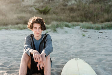 Langhaariger Teenager sitzt neben einem Surfbrett an einem Strand in Neuseeland - CAVF89532