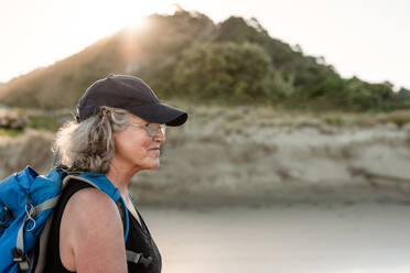 Active older woman wearing a backpack and hat walking on a beach in New Zealand - CAVF89534