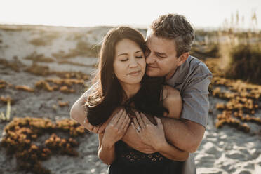 Mid-40's husband embraces beautiful wife with sand dune in background - CAVF89559