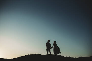 Silhouette of brother and sister standing on sand dune holding hands - CAVF89572