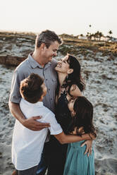 Family hug on sand dune with smiling mid-40's parents and 2 children - CAVF89576