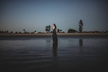 Schöne Frau in langem Kleid steht am Strand bei Sonnenuntergang - CAVF89592