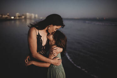 Tender mother hugging and kissing 8 yr old daughter on beach at sunset - CAVF89594