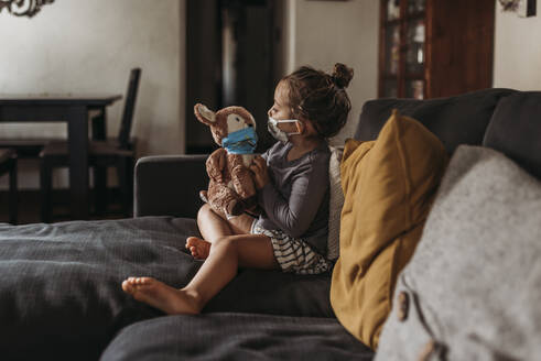 Side view of preschool age girl with mask on cuddling masked toy - CAVF89603