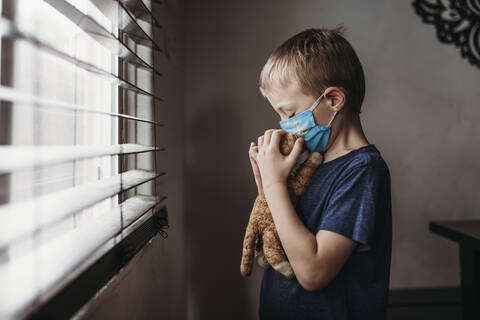 Side view of young school aged boy with mask on kissing stuffed animal stock photo