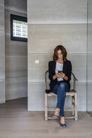 Frau, die ein Mobiltelefon benutzt, während sie auf einem Stuhl an der Wand im Büro sitzt, lizenzfreies Stockfoto
