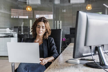 Geschäftsfrau arbeitet am Laptop, während sie auf einem Stuhl im Büro sitzt - DLTSF01239