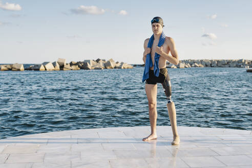 Behinderter junger Mann mit Handtuch um den Hals, der auf der Promenade am Meer steht - JCZF00240