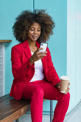 Frau hält Einweg-Kaffeebecher, während sie auf einer Bank sitzend ihr Smartphone benutzt - MGIF00997