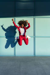 Cheerful woman with hand raised jumping against wall - MGIF01000