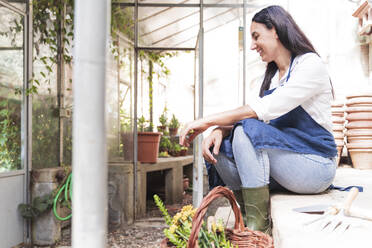 Smiling woman looking down while sitting in garden shed on sunny day - FMOF01116