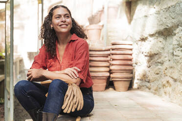 Smiling mature woman looking away with gardening glove sitting in garden shed - FMOF01119