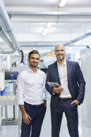 Smiling mature businessman holding digital tablet while standing by young male engineer at factory stock photo