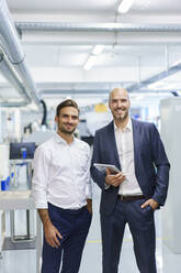 Lächelnder reifer Geschäftsmann, der ein digitales Tablet hält, während er neben einem jungen männlichen Ingenieur in einer Fabrik steht - MOEF03282