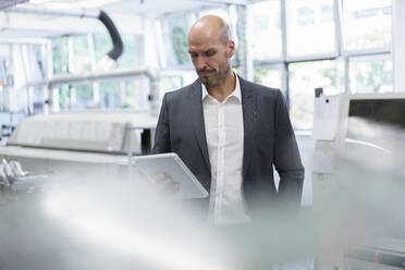 Balding businessman looking at digital tablet while standing at bright factory - MOEF03292