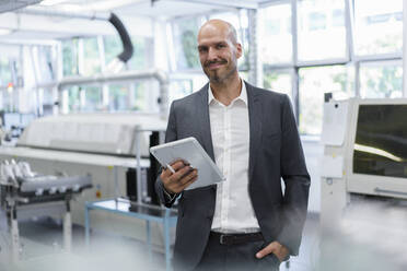 Lächelnder, selbstbewusster Geschäftsmann, der ein digitales Tablet hält, während er mit der Hand in der Tasche in einer hellen Fabrik steht - MOEF03293