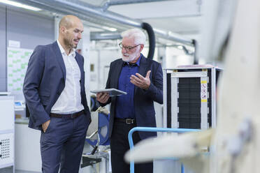 Männlicher leitender Angestellter, der mit einem Kollegen über ein digitales Tablet diskutiert, während er an einer Maschine in einer Fabrik steht - MOEF03297