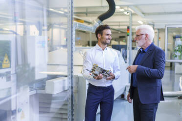 Smiling young male engineer holding machine part while discussing with senior manager at factory - MOEF03308