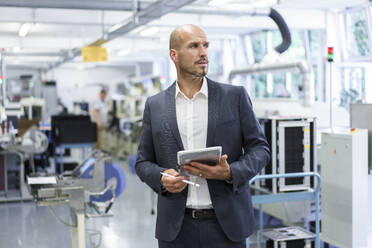 Thoughtful businessman holding digital tablet while looking away at illuminated factory - MOEF03318