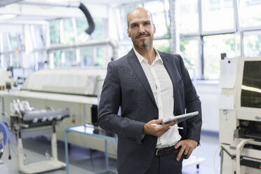 Confident businessman holding digital tablet while standing with hand on hip at illuminated factory - MOEF03320