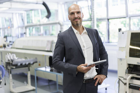 Selbstbewusster Geschäftsmann, der ein digitales Tablet hält, während er in einer beleuchteten Fabrik mit der Hand in der Hüfte steht - MOEF03320