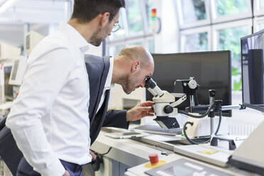 Junger männlicher Techniker steht neben einem Geschäftsmann, der durch ein Mikroskop im Labor schaut - MOEF03322