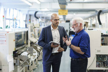 Geschäftsmann, der ein digitales Tablet hält, während er einen leitenden Angestellten in einer beleuchteten Fabrik bespricht - MOEF03327