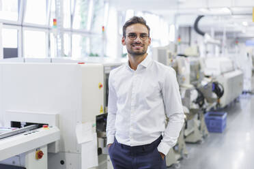 Smiling young businessman standing with hands in pockets at illuminated factory - MOEF03329