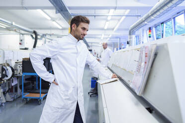 Young male technician in lab coat looking at reports on machinery - MOEF03334