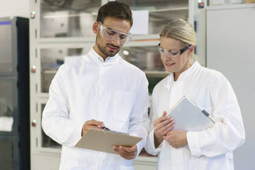 Young male technician discussing over clipboard with female blond colleague holding digital tablet at laboratory - MOEF03339