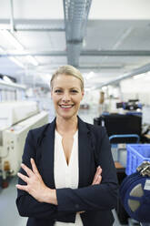 Smiling mature blond female professional standing with arms crossed at illuminated industry - MOEF03341