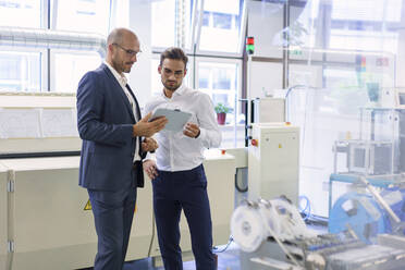 Confident male colleagues discussing over digital tablet while standing against machinery at factory - MOEF03351