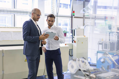 Selbstbewusste männliche Kollegen diskutieren über ein digitales Tablet, während sie in einer Fabrik vor Maschinen stehen - MOEF03351