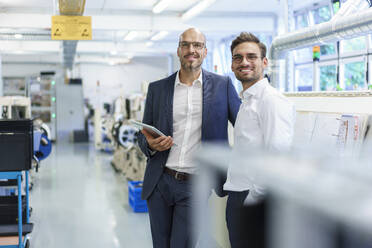 Lächelnde männliche Kollegen stehen vor Maschinen in einer beleuchteten Fabrik - MOEF03360