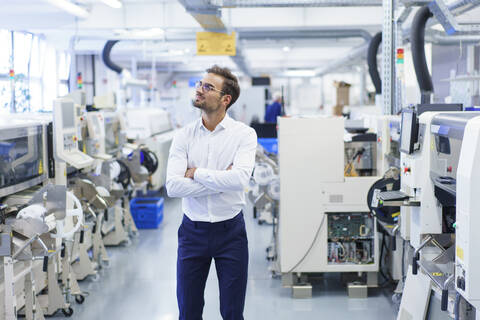 Thoughtful young businessman standing with arms crossed while looking away at illuminated factory stock photo