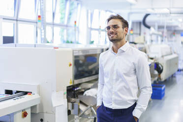 Smiling young male technician standing with hands in pockets at illuminated factory - MOEF03365