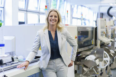 Smiling mature blond businesswoman standing by machinery at illuminated industry - MOEF03368