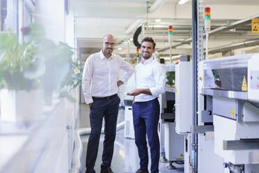 Lächelnde männliche Ingenieure, die eine Fernbedienung in der Hand halten, während sie an einer Maschine in einer Fabrik stehen - MOEF03372
