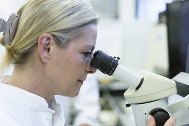 Blonde Wissenschaftlerin schaut durch ein Mikroskop in einem beleuchteten Labor - MOEF03391