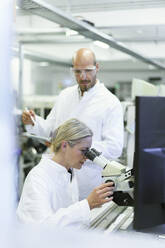 Female scientist doing research while looking into microscope by male colleague in laboratory - MOEF03392