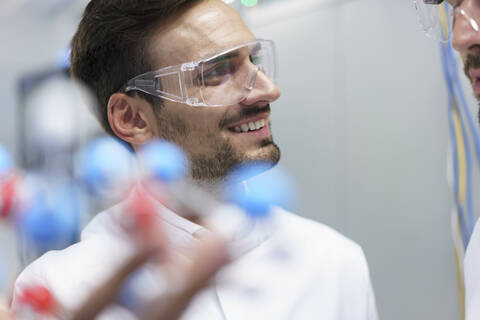 Smiling male scientist holding molecular structure while looking at colleague in illuminated laboratory stock photo