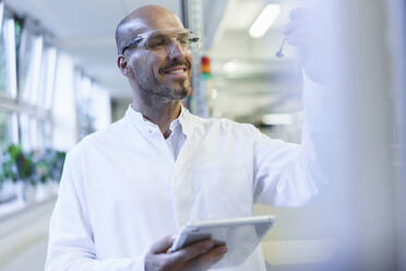 Lächelnder männlicher Wissenschaftler, der eine Probe betrachtet und ein digitales Tablet in einer Fabrik hält - MOEF03398
