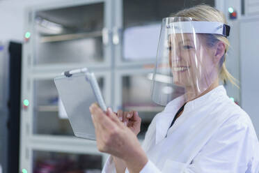 Lächelnde reife Wissenschaftlerin, die einen Gesichtsschutz trägt, während sie ein digitales Tablet im Labor benutzt - MOEF03401