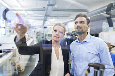 Confident businesswoman planning with young engineer while pointing at glass interface in factory - MOEF03408